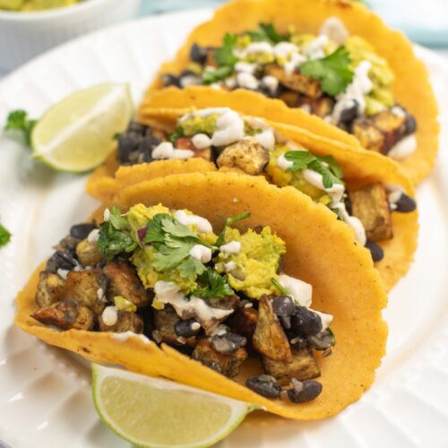 Three Sweet Potato and Black Bean Tacos filled with grilled vegetables, guacamole, cheese, and cilantro are served on a white plate with lime wedges.
