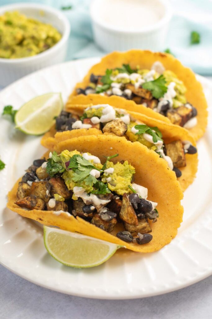 Three Sweet Potato and Black Bean Tacos filled with grilled vegetables, guacamole, cheese, and cilantro are served on a white plate with lime wedges.