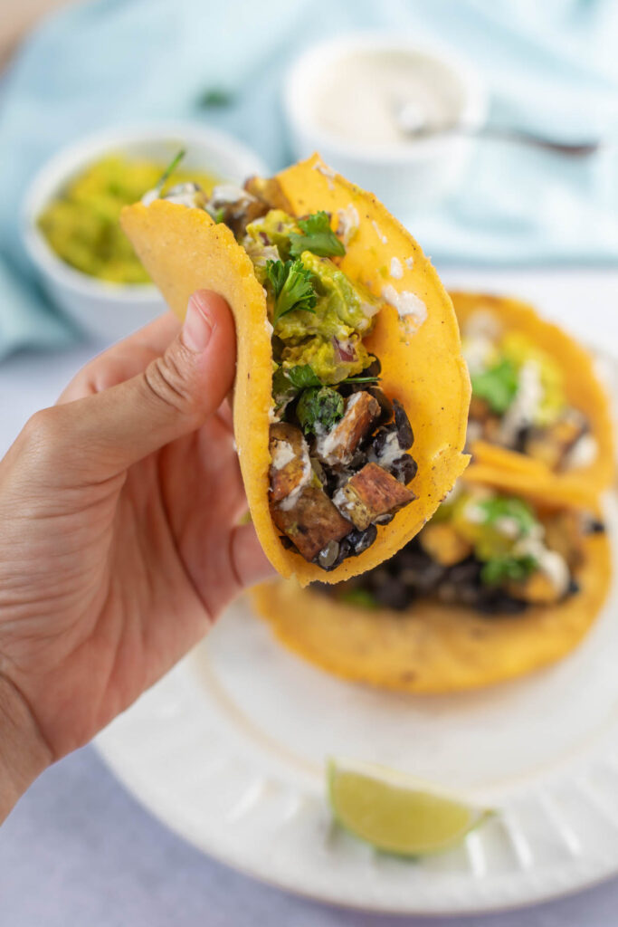 A hand holds a Sweet Potato and Black Bean Taco filled with grilled vegetables, cilantro, and sauce; more tacos and a lime wedge rest on a plate below.