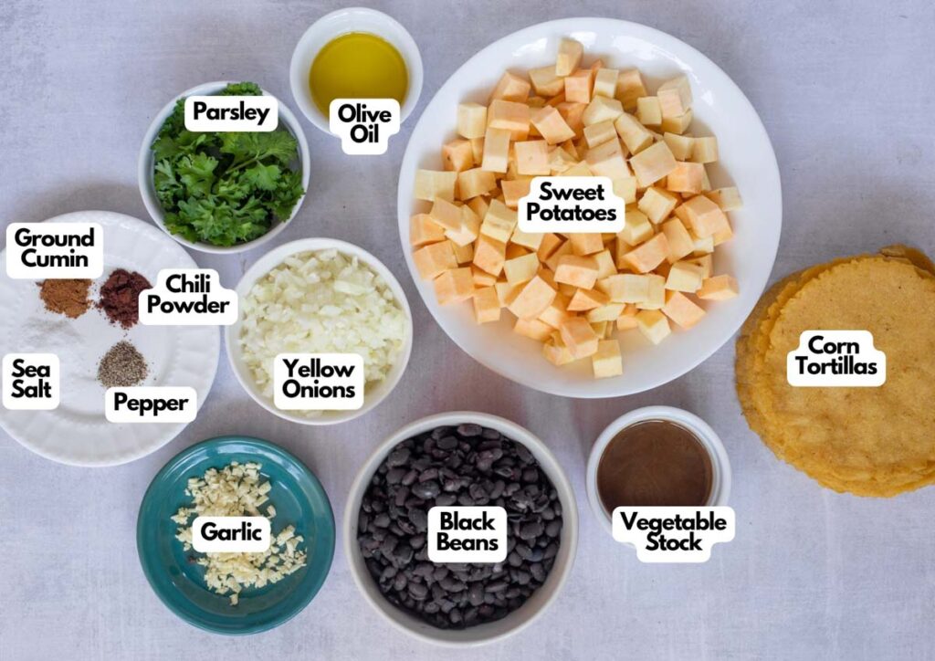 Ingredients for Sweet Potato and Black Bean Tacos are laid out in bowls and plates, including sweet potatoes, black beans, yellow onions, parsley, garlic, spices, olive oil, vegetable stock, and corn tortillas.