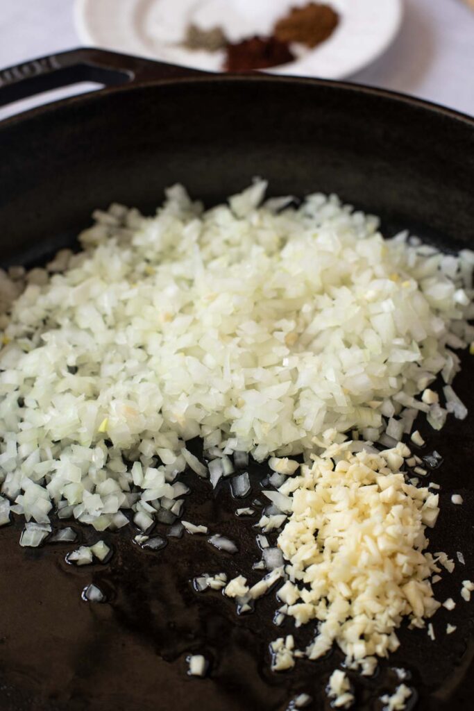 Chopped onions and minced garlic cooking in a black skillet, ready to fill flavorful Sweet Potato and Black Bean Tacos, with a plate of spices in the background.