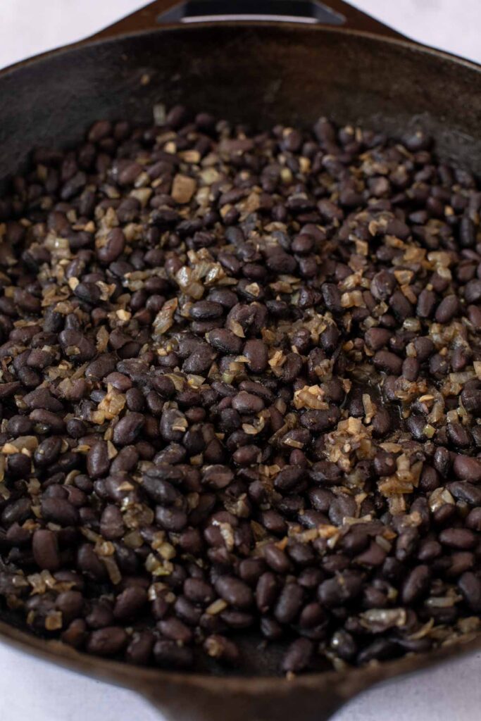 A close-up of cooked black beans mixed with diced onions in a cast iron skillet, perfect for filling delicious Sweet Potato and Black Bean Tacos.