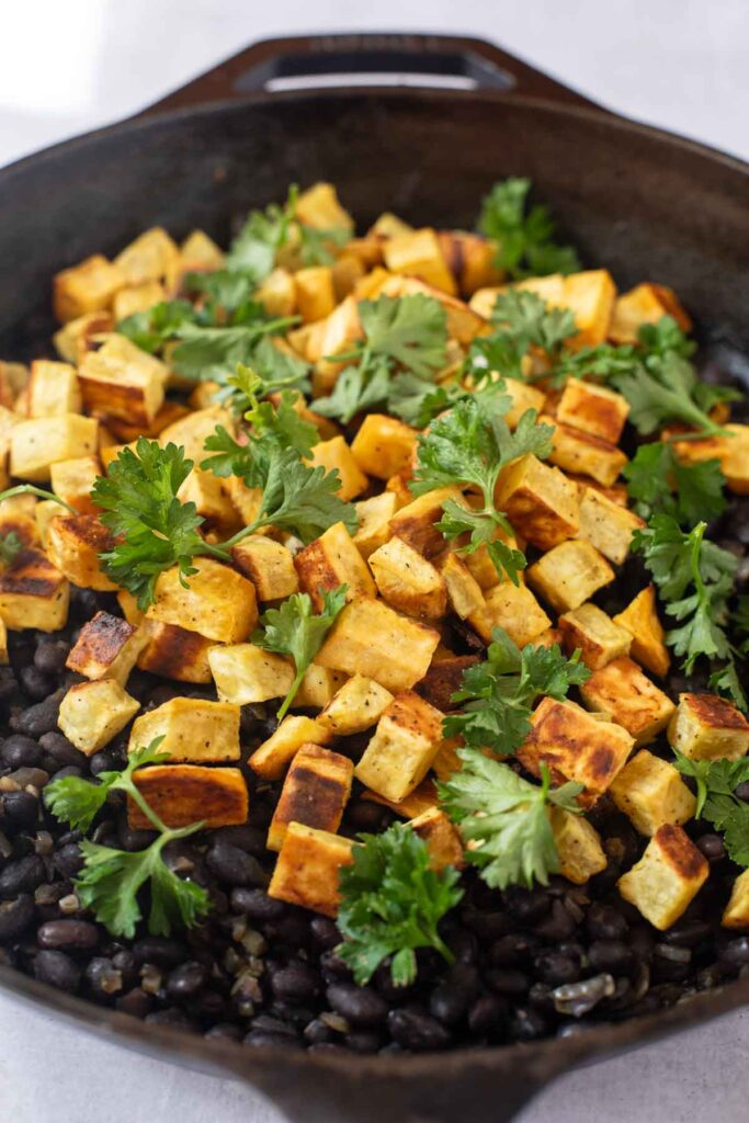 Cubed roasted tofu and fresh cilantro garnish atop cooked black beans in a cast iron skillet, perfect as a filling for Sweet Potato and Black Bean Tacos.