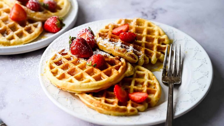 A white plate with three waffles topped with sliced strawberries and powdered sugar, with a fork beside them; another plate of waffles is in the background.