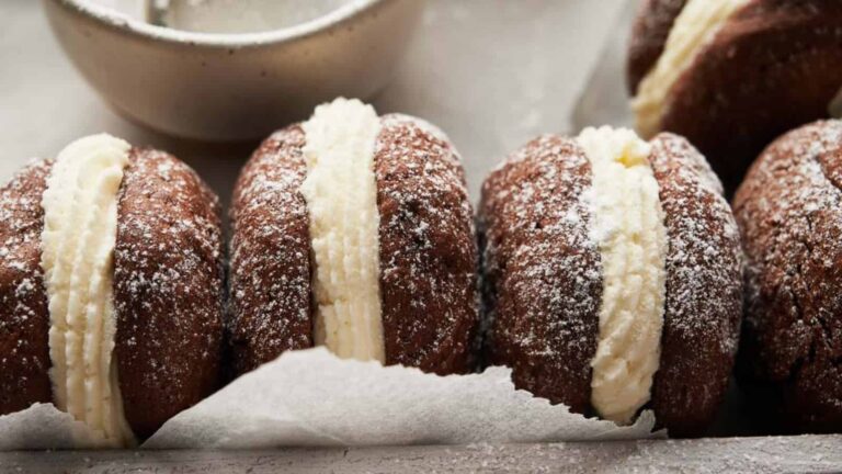 Four chocolate sandwich cookies filled with white cream and dusted with powdered sugar, lined up on parchment paper with a bowl in the background.