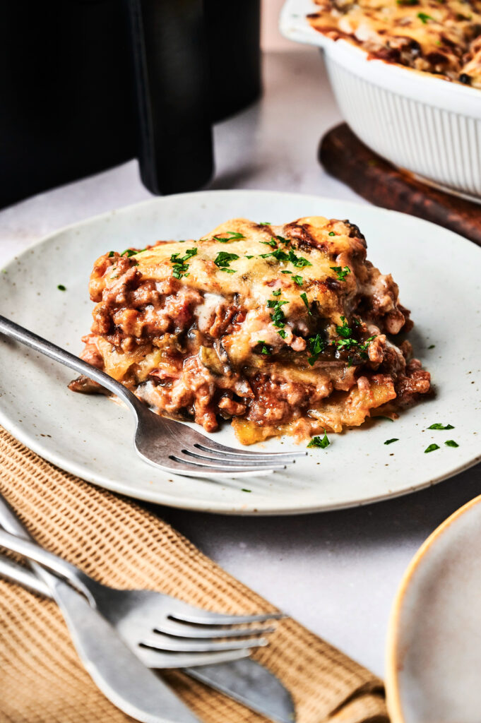 A slice of Air Fryer Beef Moussaka topped with melted cheese and chopped parsley sits on a white plate with a fork.