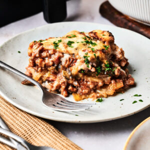 A slice of Air Fryer Beef Moussaka topped with melted cheese and chopped parsley sits on a white plate with a fork.