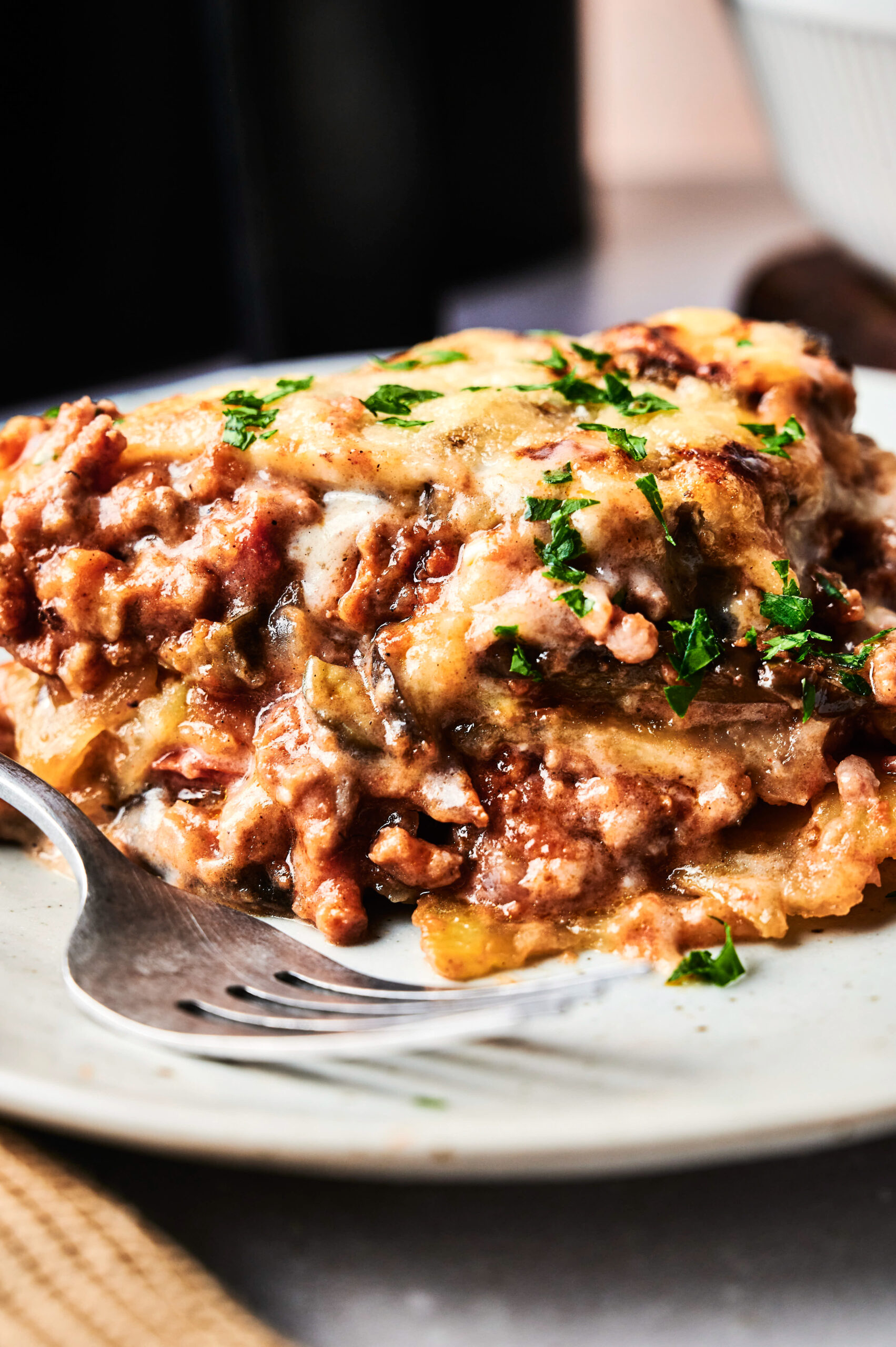A close-up of a serving of Air Fryer Beef Moussaka on a white plate, garnished with chopped parsley, with a fork beside it.