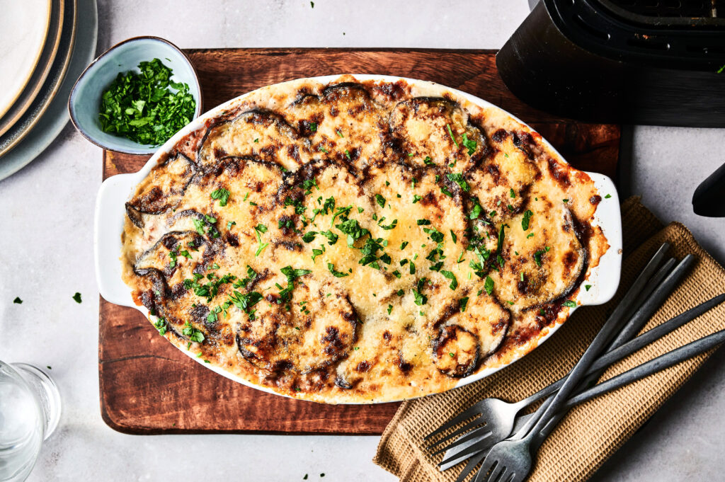 Oval dish of Air Fryer Beef Moussaka topped with melted cheese and sprinkled with chopped parsley, set on a wooden board with utensils and a bowl of herbs nearby.
