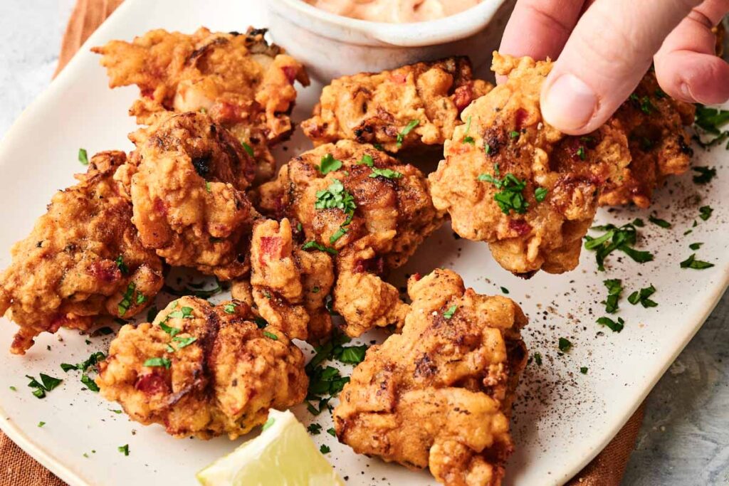 Plate of golden-brown Air Fryer Conch Fritters garnished with chopped herbs, served with a slice of lemon and a small bowl of dipping sauce; a hand holds one fritter.