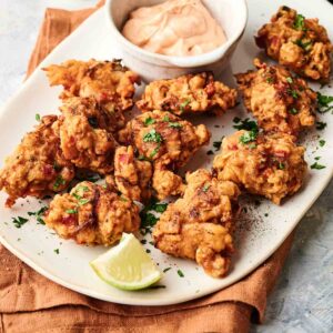 A white plate with several pieces of fried chicken and Air Fryer Conch Fritters garnished with herbs, a small bowl of dipping sauce, and lime wedges on a light surface.