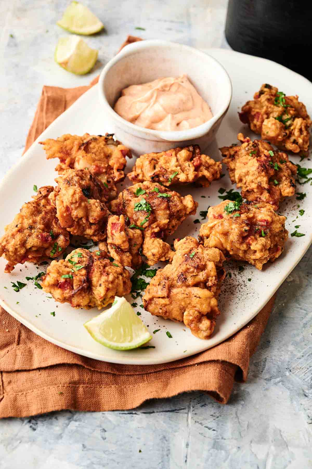 A white plate with several pieces of fried chicken and Air Fryer Conch Fritters garnished with herbs, a small bowl of dipping sauce, and lime wedges on a light surface.