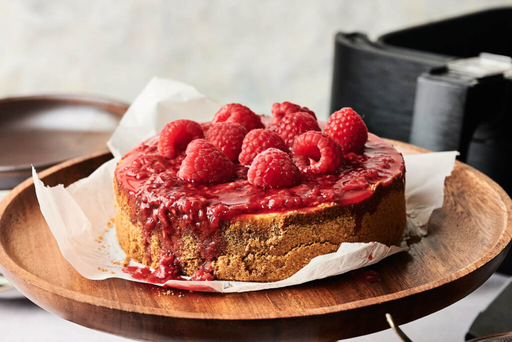 An Air Fryer Raspberry Cheesecake topped with raspberry sauce and fresh raspberries sits on parchment paper atop a wooden cake stand.
