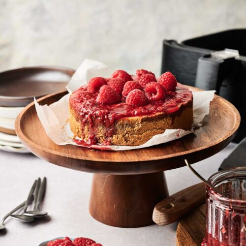 An Air Fryer Raspberry Cheesecake sits on a wooden cake stand, topped with fresh raspberries and sauce, with extra raspberries and a jar of sauce nearby.