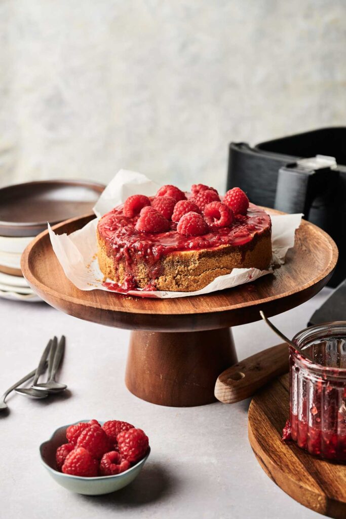 An Air Fryer Raspberry Cheesecake sits on a wooden cake stand, topped with fresh raspberries and sauce, with extra raspberries and a jar of sauce nearby.