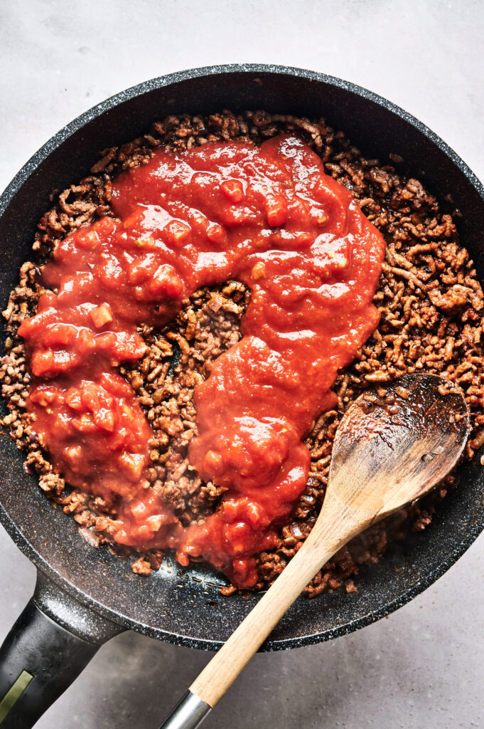 A frying pan with cooked ground beef topped with tomato sauce is being stirred with a wooden spoon, perfect for preparing the filling of an Air Fryer Beef Moussaka.