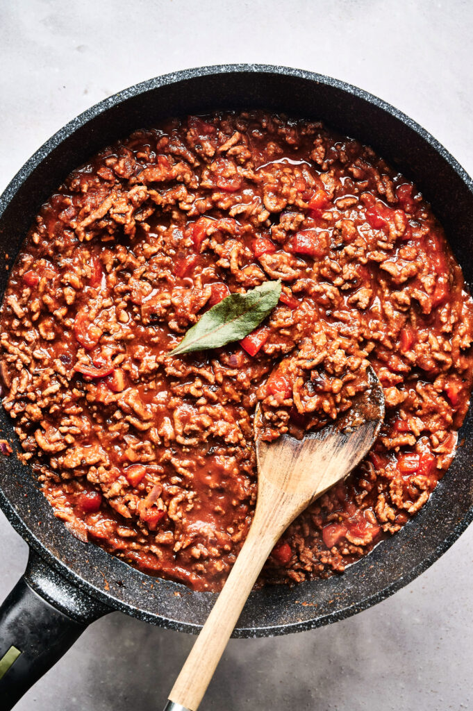 A skillet of ground meat simmered in tomato sauce with diced vegetables, reminiscent of Air Fryer Beef Moussaka, garnished with a bay leaf, and stirred with a wooden spoon.
