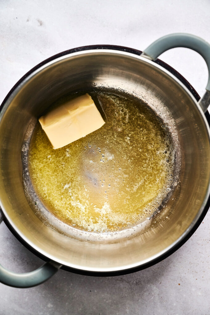 A block of butter is partially melted in a large stainless steel pot on a light-colored surface, ready to be used in dishes like Air Fryer Beef Moussaka.