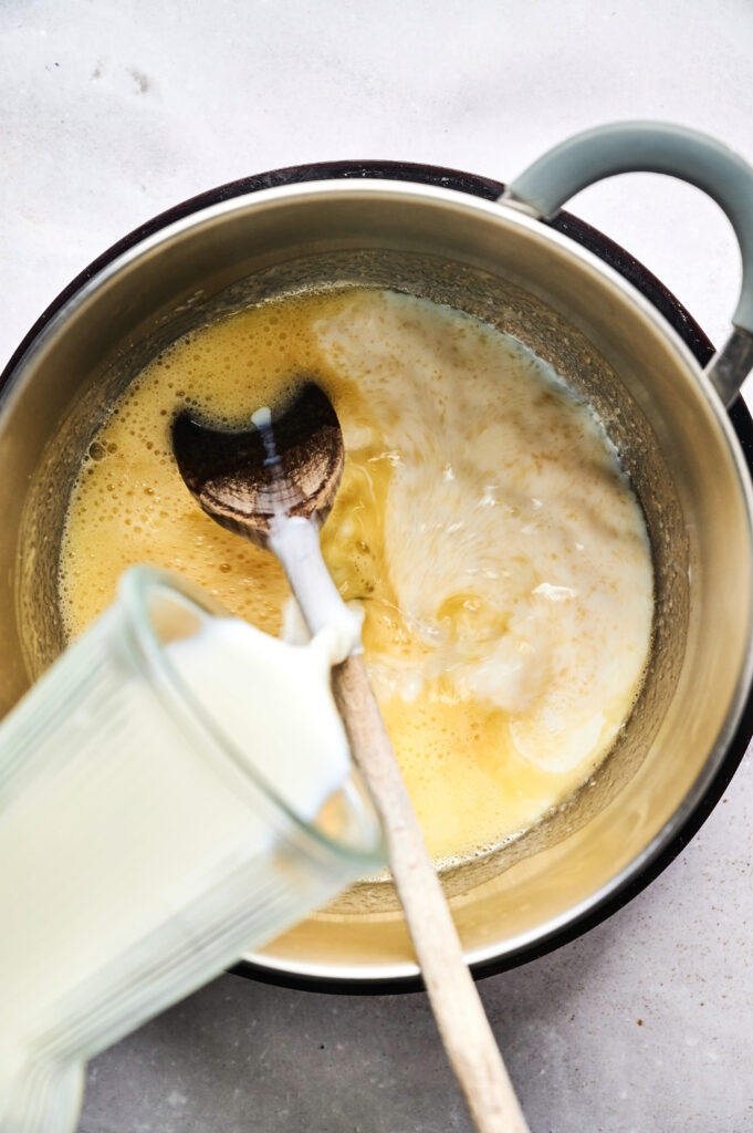 Milk is being poured from a glass into a saucepan containing beaten eggs, with a wooden spoon resting inside the pan, as part of the preparation for Air Fryer Beef Moussaka.