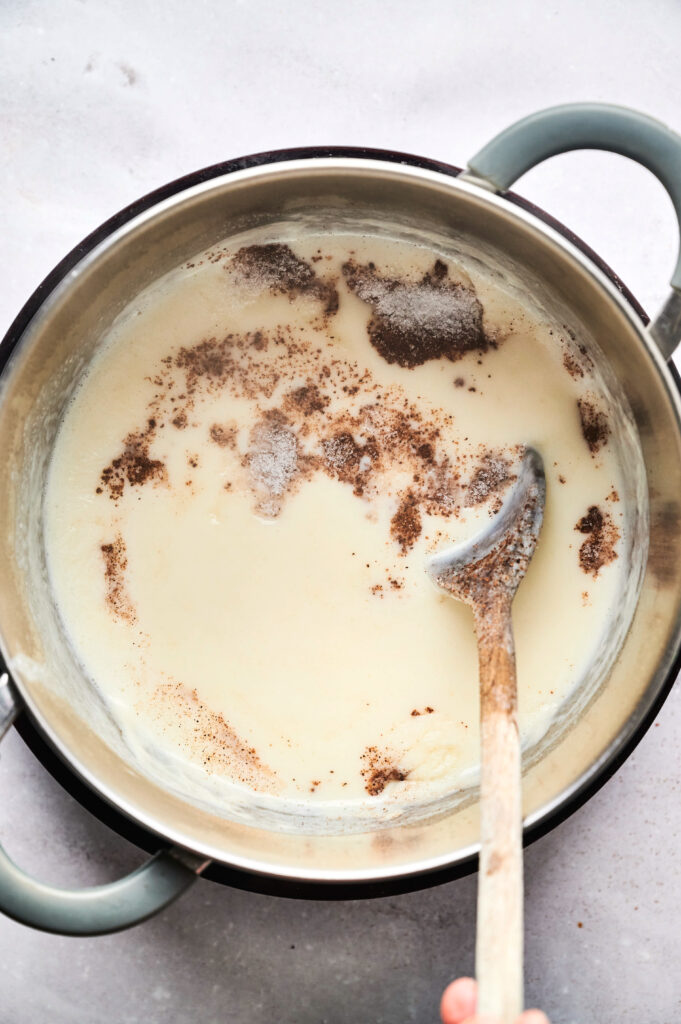 A saucepan with a light-colored sauce being stirred with a wooden spoon, topped with sprinkled brown spices&mdash;perfect for drizzling over your Air Fryer Beef Moussaka.