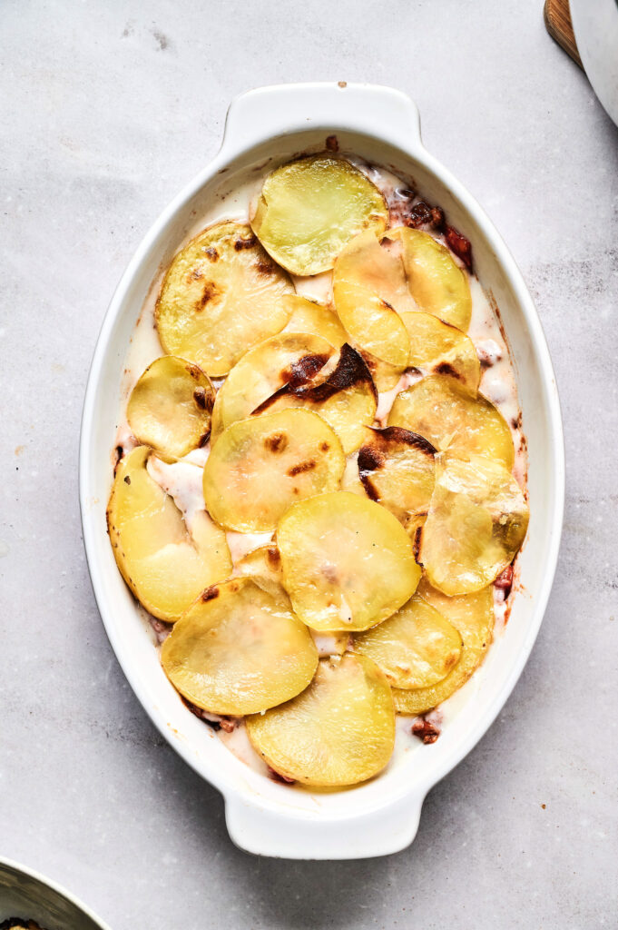 An oval white baking dish filled with layers of sliced baked potatoes in a creamy sauce, reminiscent of a classic Air Fryer Beef Moussaka, sits on a light gray surface.