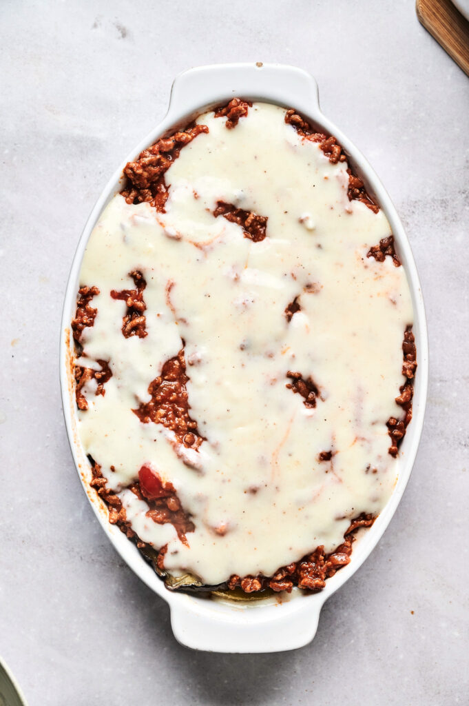 An oval baking dish filled with Air Fryer Beef Moussaka, featuring a casserole topped with a layer of white sauce over a tomato-based meat mixture, viewed from above.