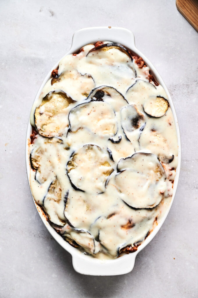 An oval baking dish filled with baked eggplant slices layered with creamy white sauce, resembling an Air Fryer Beef Moussaka, viewed from above on a light surface.
