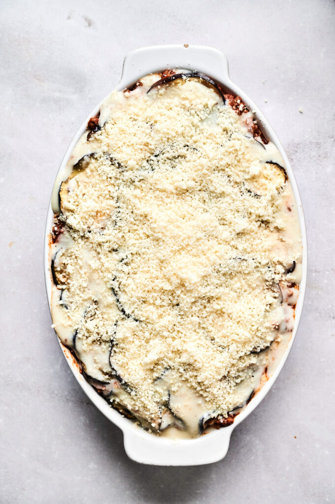 An oval baking dish filled with Air Fryer Beef Moussaka, featuring a layered casserole topped with a thick layer of grated cheese and sauce, on a light-colored surface.