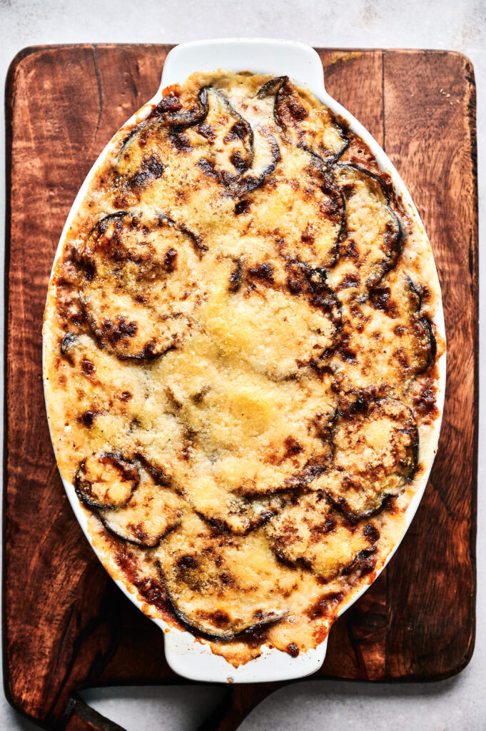 An oval baking dish filled with a golden-brown, cheesy Air Fryer Beef Moussaka casserole sits on a wooden cutting board.