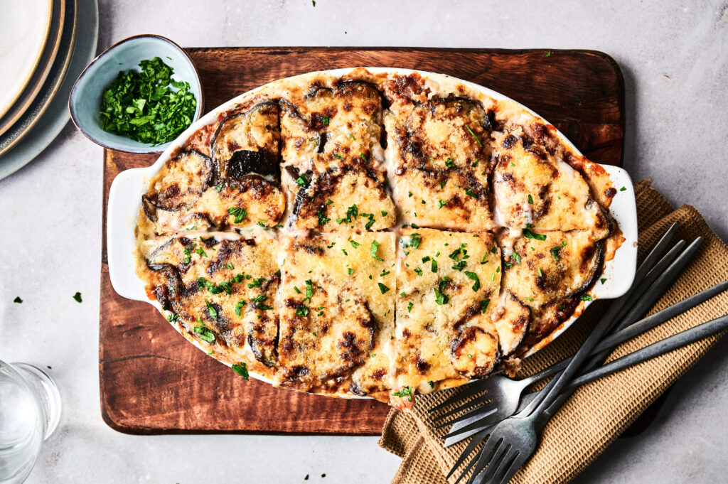 A baked dish of sliced Beef Moussaka topped with melted cheese and parsley sits on a wooden board, with a bowl of chopped herbs and utensils nearby.
