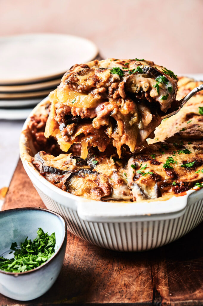 A serving of baked lasagna is lifted from a white casserole dish, with melted cheese and herbs visible; reminiscent of Beef Moussaka, a small bowl of chopped parsley sits nearby.
