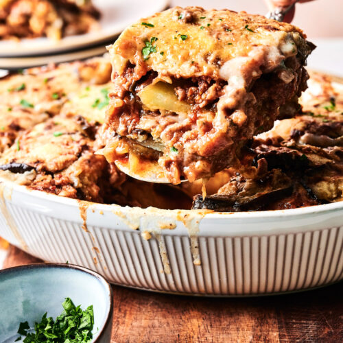 A hand serves a slice of Beef Moussaka casserole with layers of meat, potatoes, and cheese from a white dish; a bowl of chopped herbs sits nearby.