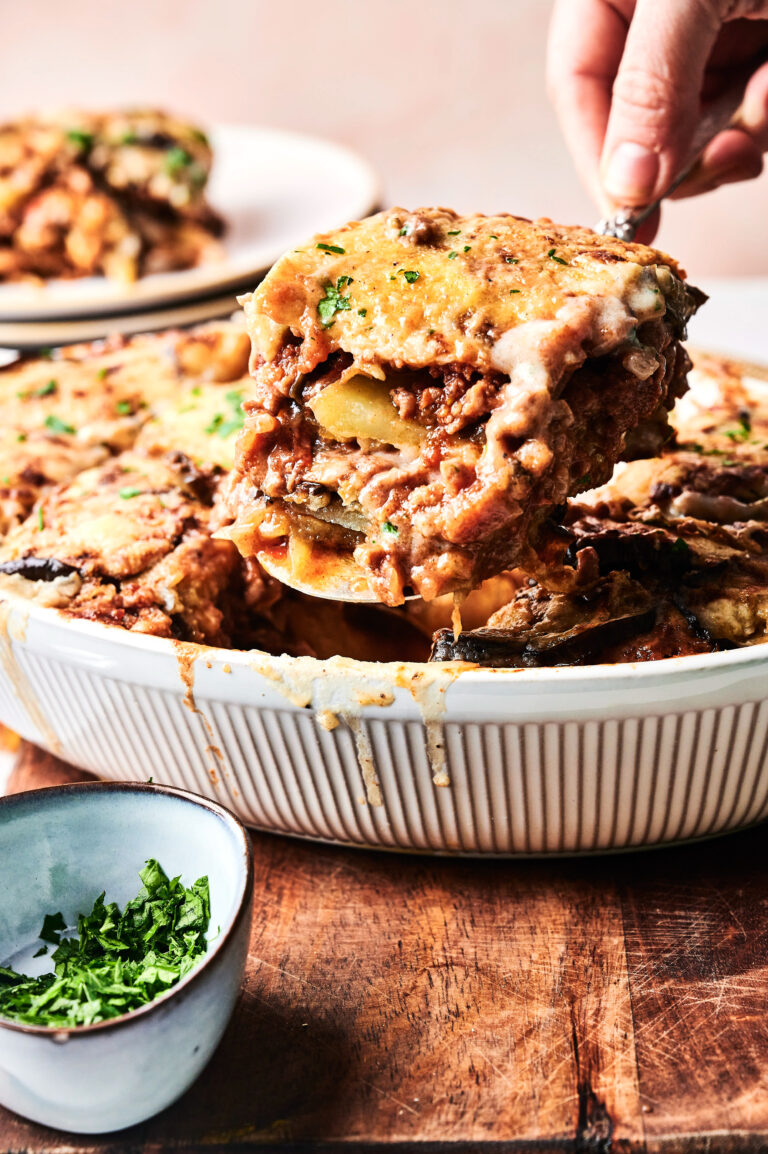 A hand serves a slice of Beef Moussaka casserole with layers of meat, potatoes, and cheese from a white dish; a bowl of chopped herbs sits nearby.