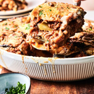 A hand serves a slice of Beef Moussaka casserole with layers of meat, potatoes, and cheese from a white dish; a bowl of chopped herbs sits nearby.