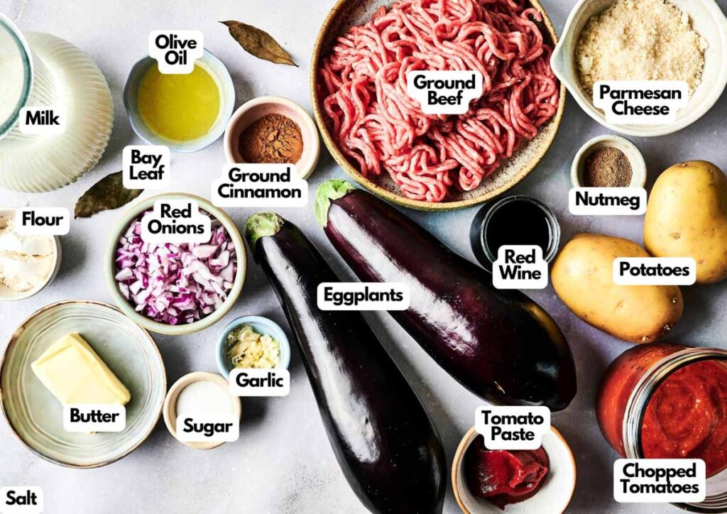 A variety of labeled ingredients for Air Fryer Beef Moussaka, including ground beef, eggplants, potatoes, red onions, milk, cheese, spices, chopped tomatoes, tomato paste, red wine, and cooking essentials.