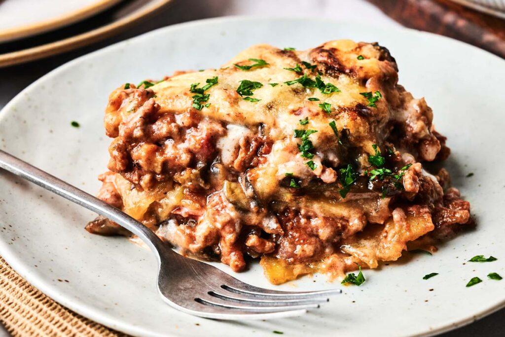 A slice of baked lasagna with meat sauce and cheese, reminiscent of Beef Moussaka, garnished with chopped parsley, served on a white plate with a fork.