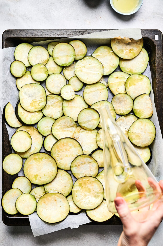 Sliced zucchini arranged on a parchment-lined baking sheet with a hand pouring oil from a glass bottle, perfect for layering in an Air Fryer Beef Moussaka.