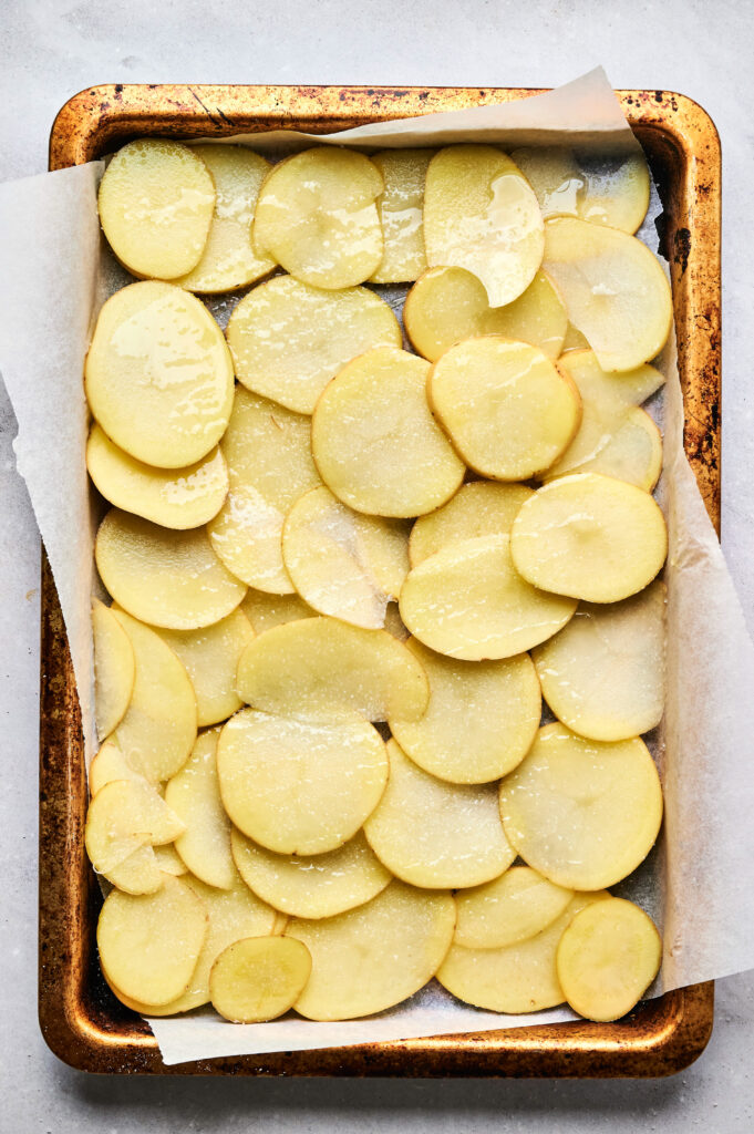 A baking tray lined with parchment paper holds a single layer of thinly sliced raw potatoes lightly coated in oil, perfect for layering in an Air Fryer Beef Moussaka.