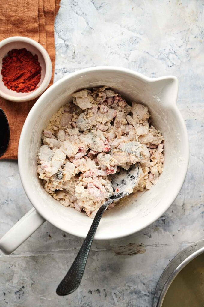 A white mixing bowl filled with a dry, chunky flour and meat mixture for Air Fryer Conch Fritters. A metal spoon rests inside the bowl, with a small bowl of red spice and an orange cloth nearby.