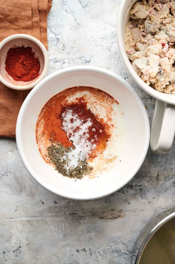 White bowl with paprika, dried herbs, salt, and pepper, next to a small bowl of paprika, a mixing bowl of Air Fryer Conch Fritters ingredients, and a tan napkin on a light surface.