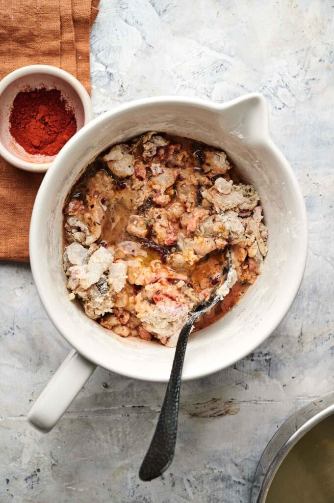 A white mixing bowl with flour, liquid, and bacon bits being stirred with a metal spoon&mdash;perfect for making Air Fryer Conch Fritters; a small bowl of paprika sits nearby on an orange cloth.