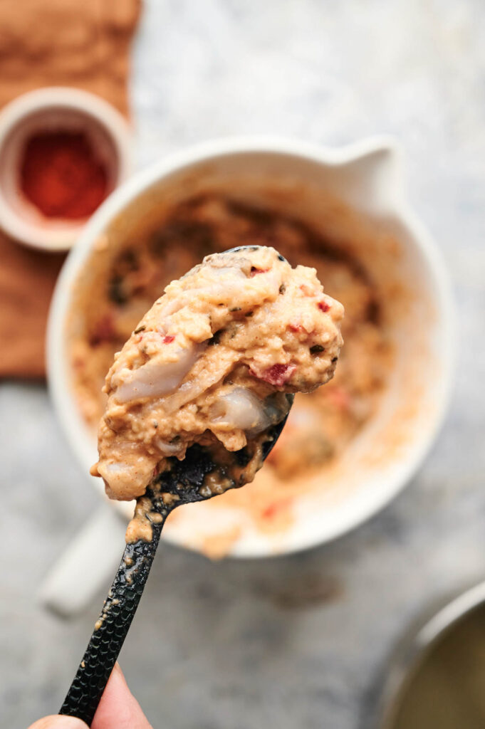 A spoonful of thick, beige batter for Air Fryer Conch Fritters, with visible pieces of onion and tomato, is held above a mixing bowl filled with more of the same savory mixture.