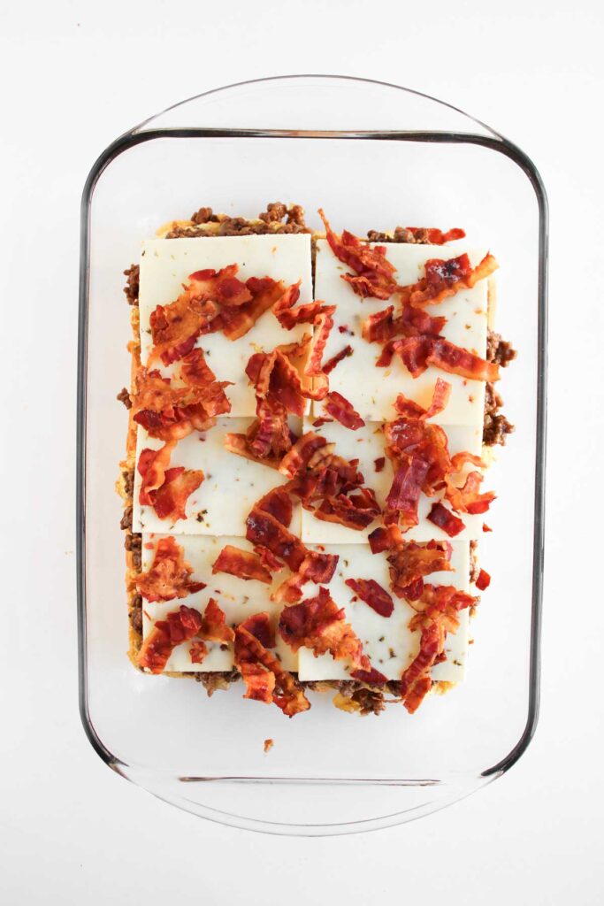A glass baking dish contains a layered casserole topped with white cheese slices and crumbled cooked bacon, reminiscent of the bold flavors found in Cowboy Sliders.