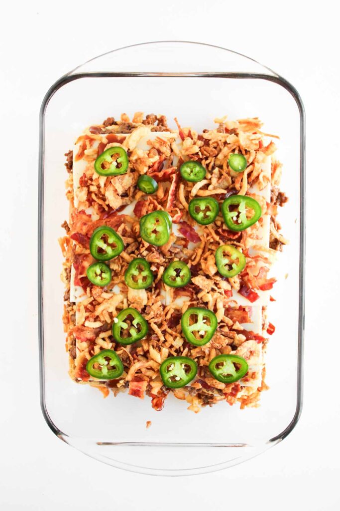 A glass baking dish holds a layered casserole inspired by Cowboy Sliders, topped with ground meat, shredded cheese, fried onions, and sliced jalape&ntilde;os.