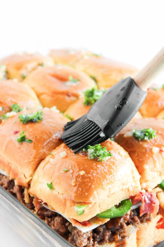 A silicone brush applies melted butter and herbs to a tray of Cowboy Sliders, featuring beef and cheese, inside a glass baking dish.
