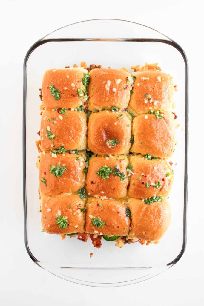 A glass baking dish containing twelve golden-brown Cowboy Sliders rolls topped with chopped herbs and melted cheese, viewed from above.
