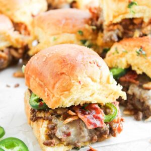 A close-up of Cowboy Sliders: mini sandwiches filled with ground meat, melted cheese, bacon, and sliced jalapeños on a white surface.