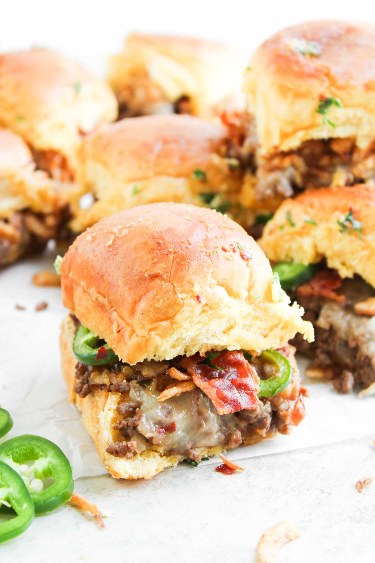 A close-up of Cowboy Sliders: mini sandwiches filled with ground meat, melted cheese, bacon, and sliced jalape&ntilde;os on a white surface.