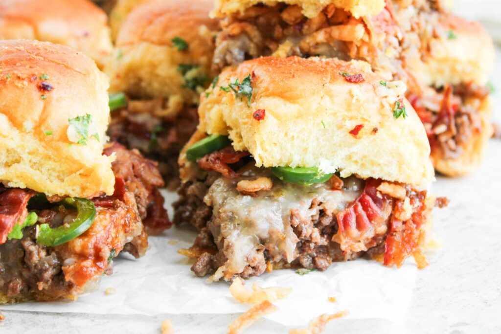 Close-up of Cowboy Sliders: cheesy beef sliders piled high with bacon, jalape&ntilde;os, and toasted buns, arranged on parchment paper.