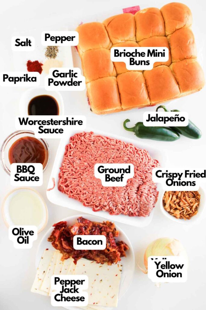 Ingredients for Cowboy Sliders are arranged on a white surface, featuring ground beef, brioche mini buns, sauces, spices, bacon, cheese, jalape&ntilde;o, crispy fried onions, and a yellow onion.